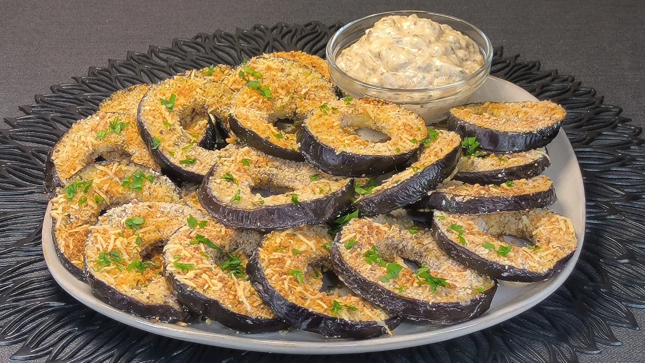 Aros de berenjena al horno rebozados y crujientes, servidos en un plato blanco acompañados de un cuenco pequeño con salsa de tomate casera sobre una mesa de cocina.