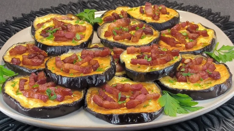 Rodajas de berenjena asadas cubiertas con salsa carbonara cremosa, bacon crujiente y queso fundido, presentadas en una bandeja de horno sobre papel vegetal.