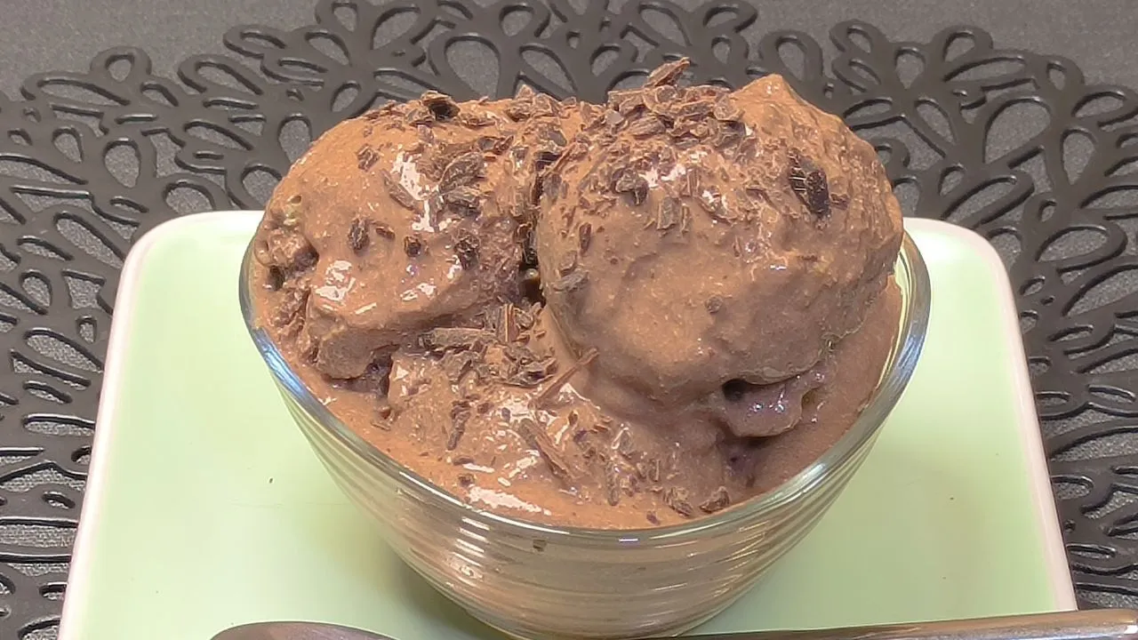 Helado cremoso de chocolate hecho con queso cottage servido en una taza de cerámica beige con trozos de almendra por encima y una cuchara de madera.