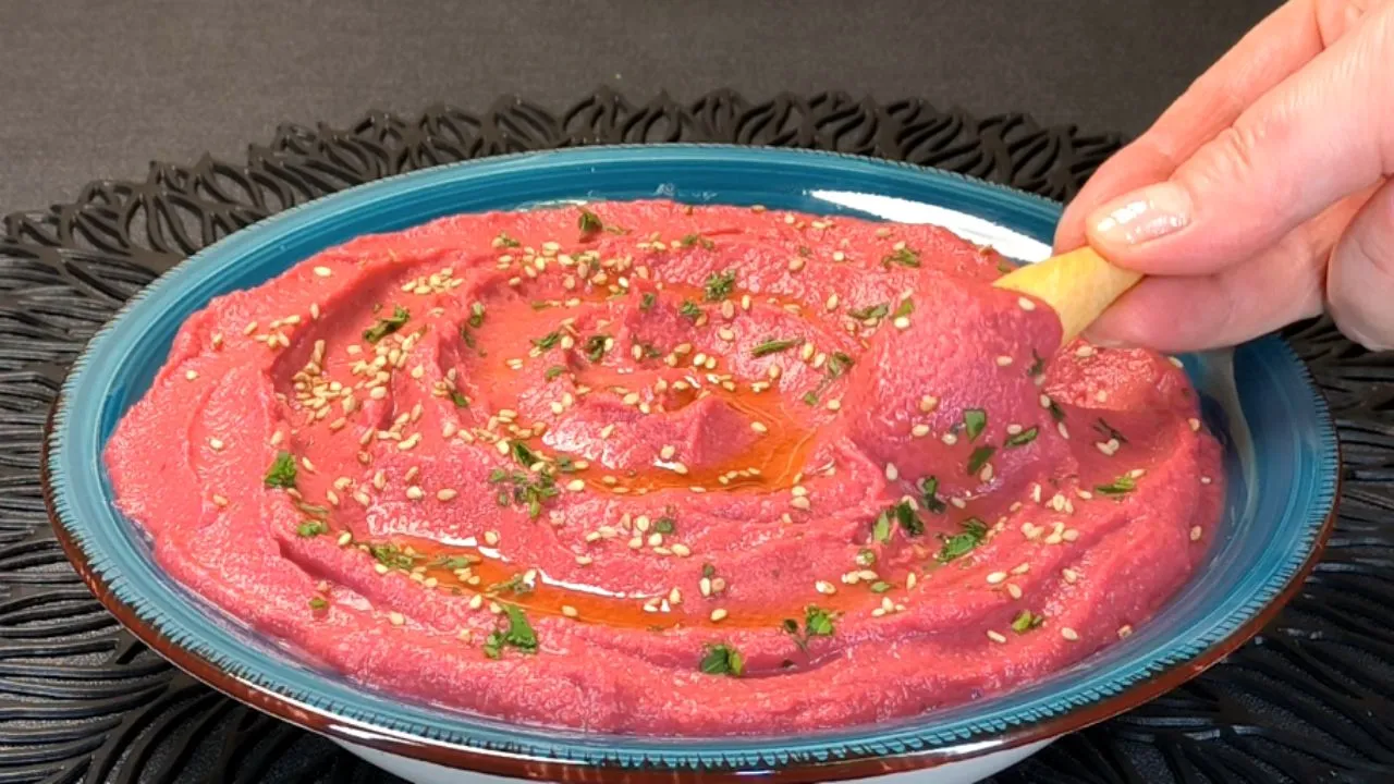 Hummus de remolacha y garbanzos de color rosa vibrante servido en un bol, decorado con un chorrito de aceite de oliva, semillas y garbanzos enteros.