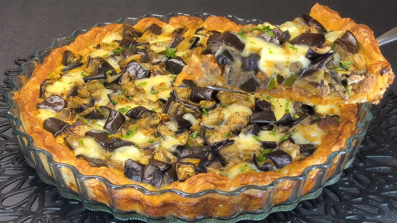 Corte de pastel de hojaldre dorado relleno de capas de berenjena, carne picada y queso fundido.
