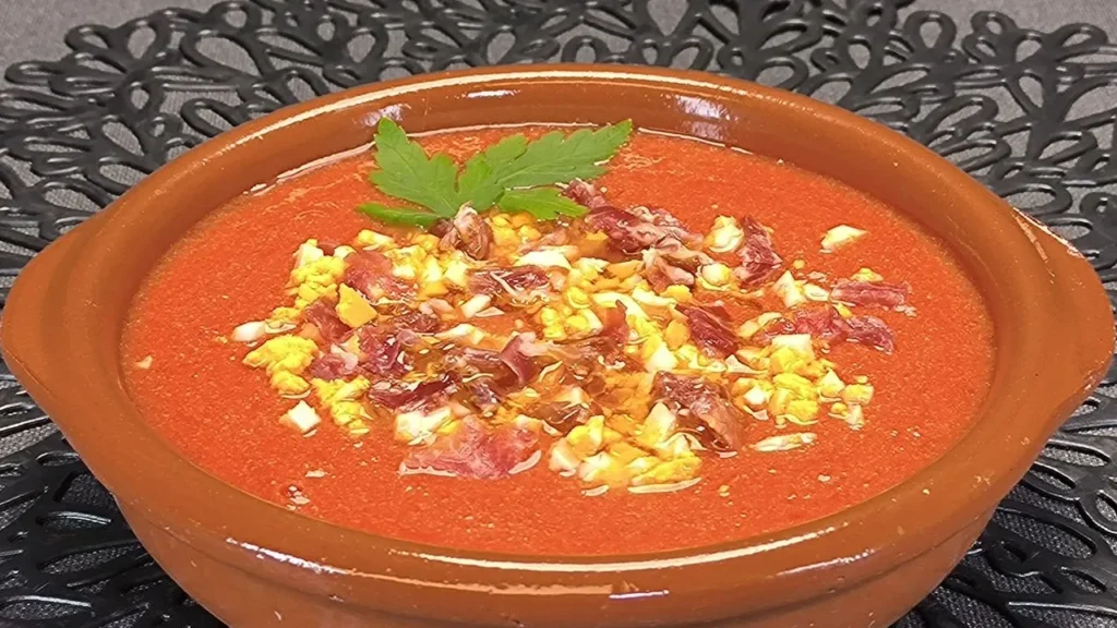 Un cuenco de barro con salmorejo cordobés cremoso, decorado con huevo duro picado y taquitos de jamón serrano, servido con un chorrito de aceite de oliva virgen extra.