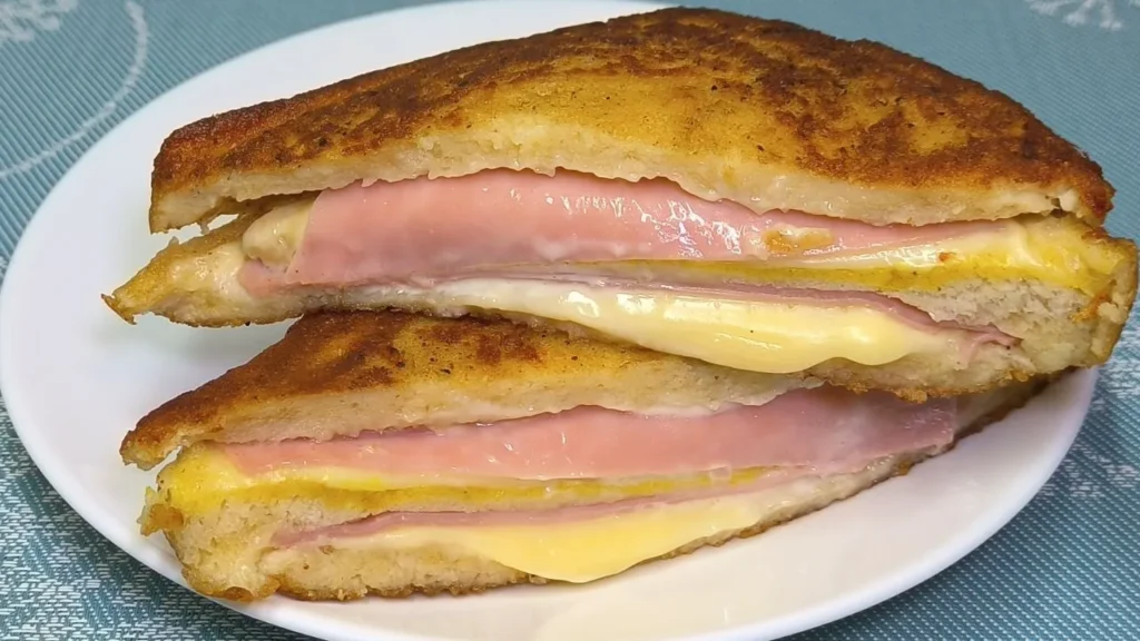 Sándwich Monte Cristo partido a la mitad mostrando capas de jamón, queso fundido y pan rebozado dorado sobre un plato.
