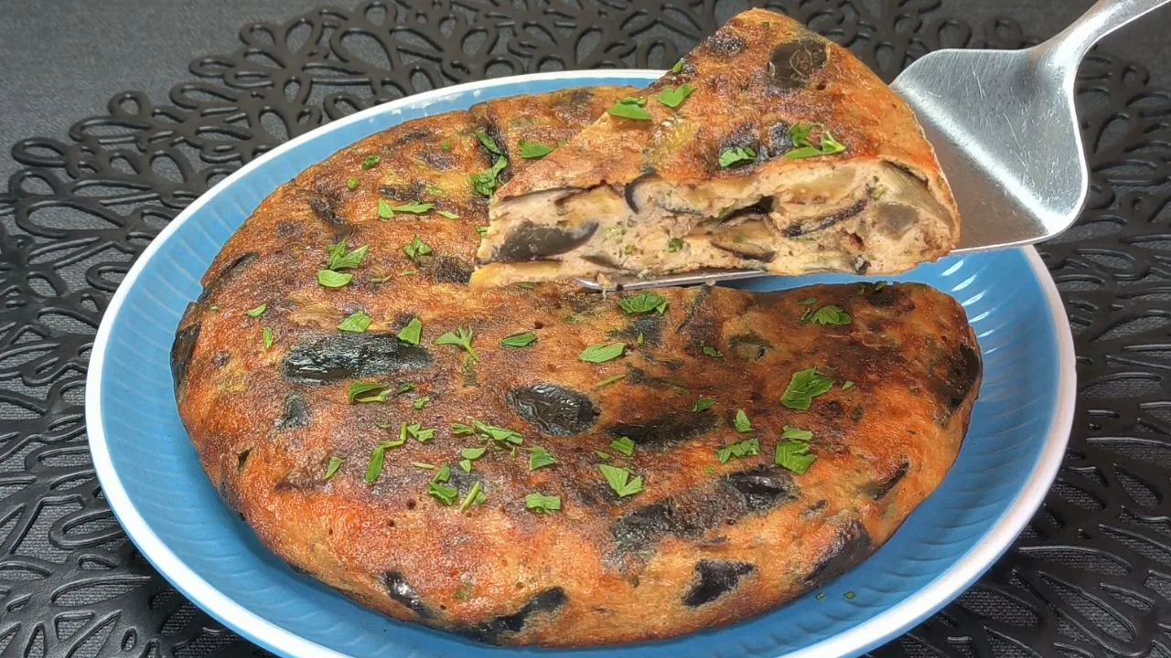 Tortilla española de berenjena con trozos de queso parmesano fundido servida en un plato blanco sobre una mesa de madera.