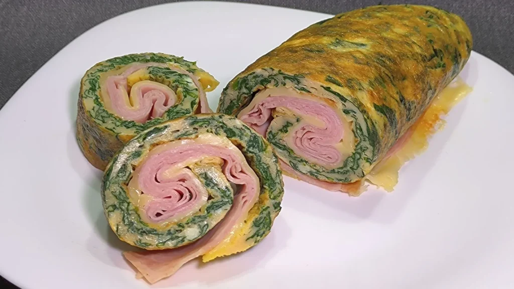 Tortilla de espinacas enrollada de color verde vibrante, rellena de queso crema y jamón, cortada en rodajas sobre un plato.
