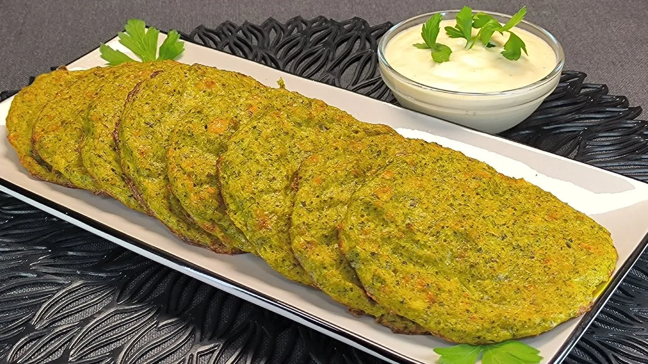 Tortitas de brócoli al horno doradas servidas en una tabla de madera con un cuenco de salsa blanca y ramitas de perejil fresco.